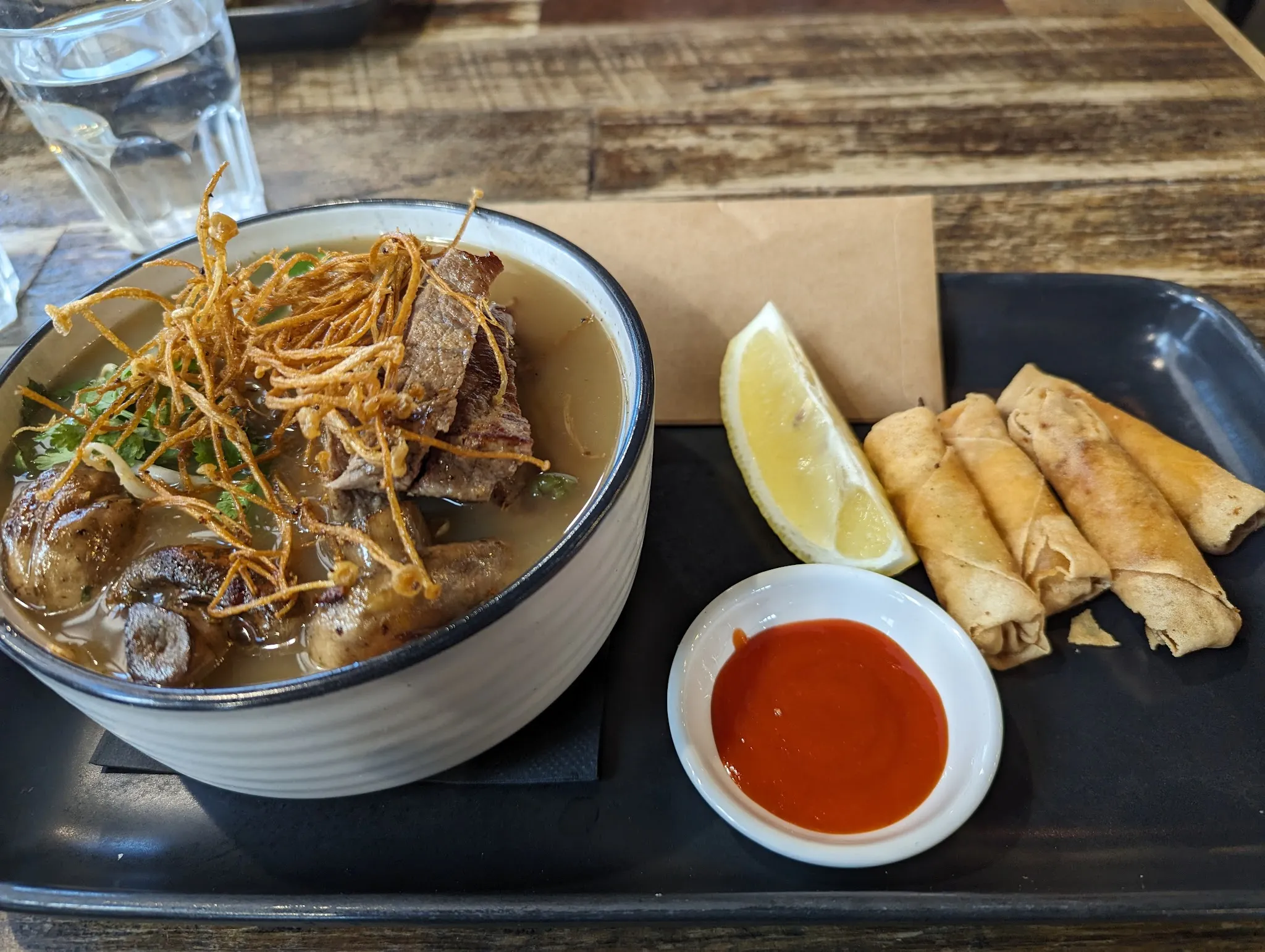 Asian-style braised pork noodle soup with spring rolls and chilli sauce on dark tray
